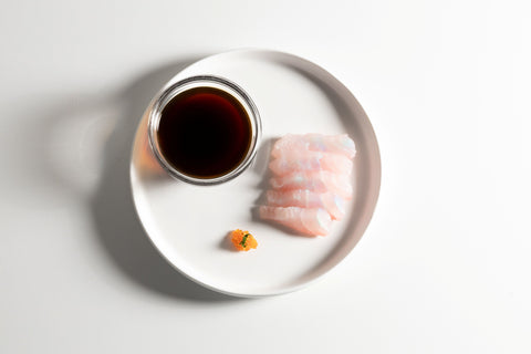 Raw fish on a white plate with a small bowl of soy sauce on a light gray background
