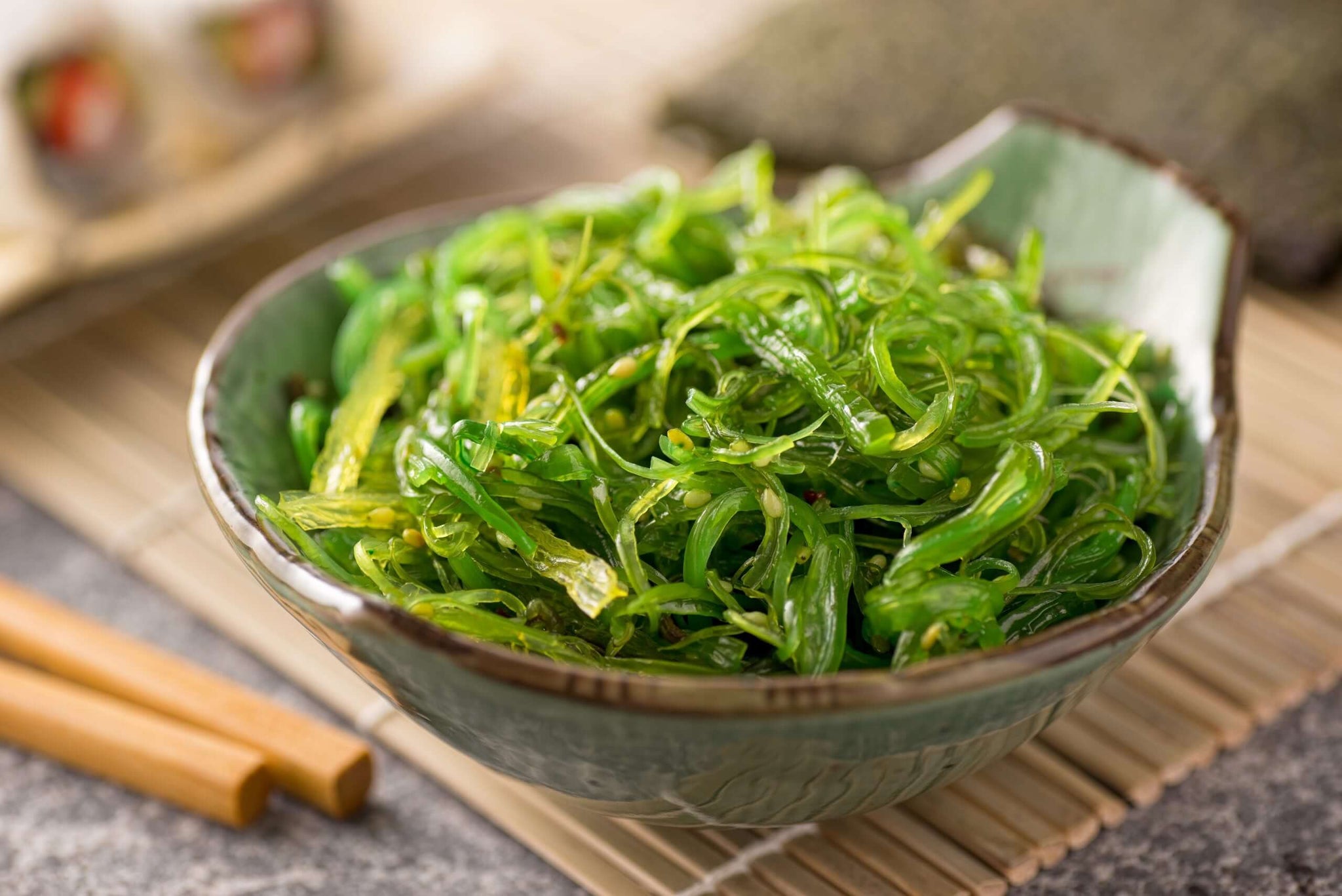 The image showcases a vibrant and appetizing serving of seaweed salad, presented in a uniquely shaped, light green ceramic bowl with a dark brown rim. The salad itself is a lush, tangled mass of bright green seaweed strands, appearing fresh and slightly glistening, suggesting a light dressing. Scattered throughout the seaweed are tiny, darker green specks, likely sesame seeds, adding texture and visual interest. The bowl is positioned on a rectangular bamboo placemat, providing a natural and earthy backdrop. To the left of the bowl lie a pair of light-colored wooden chopsticks, ready for enjoying the salad. In the blurred background, partially visible are slices of sushi rolls, hinting at a Japanese culinary theme. A sheet of nori seaweed is also subtly present in the background, further reinforcing this association. The surface beneath the placemat appears to be a textured, grey stone or concrete, offering a contrasting cool tone to the warm bamboo and vibrant green of the salad. The lighting is soft and diffused, highlighting the textures and colors of the food and surrounding elements, creating an inviting and appealing composition. The overall impression is one of fresh, healthy, and flavorful Japanese cuisine.